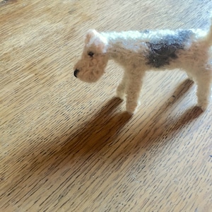 May include: A white and black felt wirehaired fox terrier dog figurine. The dog is standing on a wooden surface.
