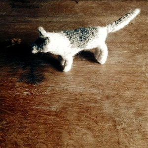 May include: A small knitted grey and white dog toy. The toy is lying on a wooden surface.