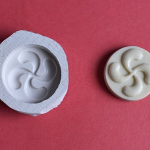 Peut inclure: Un moule circulaire blanc et un objet circulaire crème assorti avec un motif stylisé en quatre parties, sur fond rouge. Le moule a un bord rugueux et irrégulier, tandis que l'objet est lisse et plein.