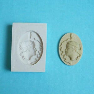 Op de afbeelding: Een witte rechthoekige mal en een beige ovale cameo, beide met een profiel van een vrouw met een hoed. De cameo is een afgewerkt product, terwijl de mal bedoeld is om vergelijkbare items te creëren. De achtergrond is effen lichtblauw.