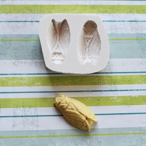 May include: A white silicone mold with two cicada wing impressions and a tan cicada. The mold is rectangular and sits on a striped background of white, green, and blue. The cicada is a detailed insect replica.
