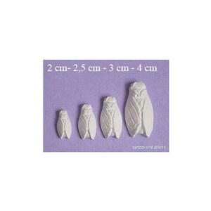 May include: Four white cicada-shaped decorations of varying sizes, ranging from 2 cm to 4 cm in length, are displayed on a lavender background. The decorations have detailed wing and body patterns. The text "2 cm - 2,5 cm - 3 cm - 4 cm" is at the top.