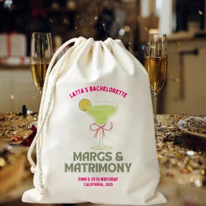 May include: A cream-coloured drawstring bag featuring the text "Latta's Bachelorette" in pink, and a margarita graphic. Below the graphic, the text "MARGS & MATRIMONY" and "FINN'S 25TH BIRTHDAY CALIFORNIA, 2025" are printed in green. Two champagne flutes are in the background.