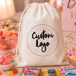 Könnte beinhalten: Ein cremefarbener Stoffbeutel mit Kordelzug, der eine schwarze kreisförmige Grafik mit dem Text Custom Logo in der Mitte aufweist. Der Beutel besteht aus einem weichen Textilmaterial und hat zwei weiße geflochtene Kordeln zum Zuziehen.