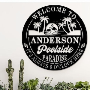 May include: Black and white metal sign with the words "WELCOME TO" at the top. The sign features palm trees, flamingos, and the text "ANDERSON Poolside PARADISE". The phrase "IT'S ALWAYS 5 O'CLOCK HERE" is also included.