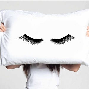 May include: White pillow featuring a black eyelash design. The pillow displays two sets of closed eyelashes, offering a simple yet stylish aesthetic. The design is centered, providing a clean and modern appearance.