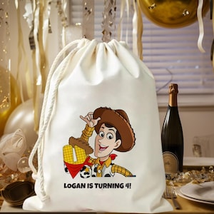 May include: A white drawstring bag featuring a cartoon cowboy character with a brown hat, red neckerchief, and yellow plaid shirt. The text "LOGAN IS TURNING 4!" is printed below the character. The bag is tied with a white rope.