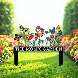 May include: A black metal garden sign with the text "THE MOM'S GARDEN". The sign features colorful butterflies and flowers. The sign is placed in a garden with green grass and colorful flowers.