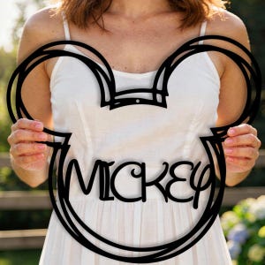 Könnte beinhalten: Schwarze Metall-Silhouette eines Mickey Mouse-Kopfes mit dem Namen "Mickey" in kursiver Schrift ausgeschnitten. Das Kunstwerk wird vor einem weißen Kleid und einem verschwommenen Hintergrund im Freien gehalten.