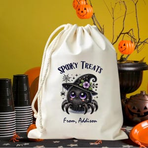 May include: A cream-colored drawstring bag with the words "Spooky Treats" and "From, Addison" printed on it. The bag features a cartoon spider wearing a witch hat with purple flowers. The spider has large purple eyes and is surrounded by a spiderweb.