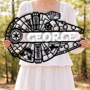 May include: Black metal wall art in the shape of the Millennium Falcon, with the name "GEORGE" in white. The design features Star Wars elements such as the Death Star and Darth Vader's helmet. The art is held against a blurred green backdrop.