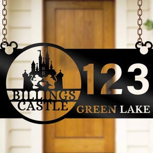 Könnte beinhalten: Schwarzes Metall-Adressschild mit einer Schloss-Silhouette, Mickey Mouse-Ohren und den Worten "Billings Castle Green Lake" mit der Nummer "123". Das Schild hängt an Ketten.