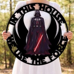 Könnte beinhalten: Runder schwarzer Metallschild mit Darth Vader und einem roten Lichtschwert. Der Schriftzug "IN THIS HOUSE WE USE THE FORCE" ist weiß. Der Hintergrund ist ein verschwommenes Bild eines Waldes.