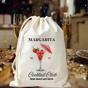 May include: A cream-coloured drawstring bag featuring the word "MARGARITA" in black. It displays a watercolour illustration of a margarita cocktail with strawberries and a red umbrella. The bag also has the text "Cocktail Club" and "DRINK MARGS SAVE WATER".