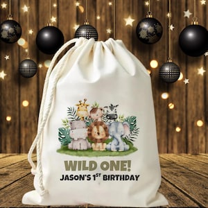 Può includere: Una borsa con coulisse color crema con un'illustrazione colorata di animali safari dei cartoni animati, tra cui una giraffa, un leone, una zebra e un elefante. Il testo "WILD ONE!" e "JASON'S 1ST BIRTHDAY" sono stampati sotto. Ornamenti neri sono appesi sullo sfondo.
