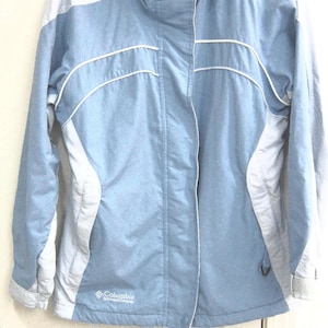 May include: A light blue and gray Columbia Sportswear Company hooded jacket. The jacket has a zipper closure, white piping details, and a drawstring hood. The jacket is hanging on a white hanger.