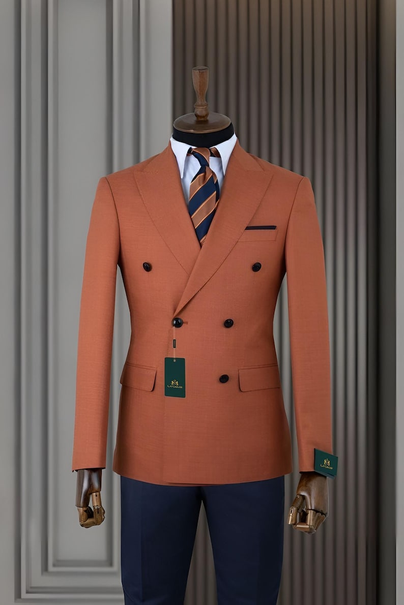 Pode incluir: Casaco de fato cor de ferrugem de abotoamento duplo com bot&otilde;es pretos, gravata listrada azul marinho e laranja e camisa branca. O fato &eacute; exibido em um manequim com cal&ccedil;as azul marinho.