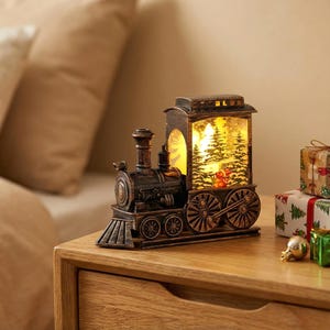 May include: A bronze-coloured Christmas train ornament with a snow globe feature. The train has intricate detailing and a snow globe section showing a winter scene with trees and a figure. It is displayed on a wooden surface.