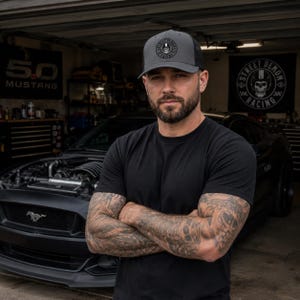 May include: A gray and black trucker hat with the text "STREET DEMON RACING" and a skull logo. The hat is worn by a person wearing a black t-shirt. A black Mustang car is in the background.
