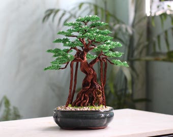 Puede incluir: Una escultura de árbol bonsái hecha a mano con alambre, con un tronco retorcido de color cobre y raíces aéreas, con follaje de cuentas verde vibrante dispuesto en capas, todo en una maceta ovalada de cerámica gris oscuro con piedras.