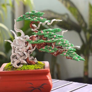 May include: A handcrafted wire bonsai tree sculpture. The tree features a silver wire trunk, green leaves, and small pink flowers. It is planted in a red rectangular pot with a moss base, creating a miniature garden scene.