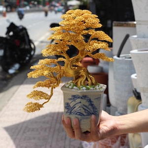 May include: A golden wire bonsai tree in a small, decorative ceramic pot with a blue and white landscape design. The bonsai has detailed leaves and branches, and is planted with small white stones. The pot is held in front of a blurred background.