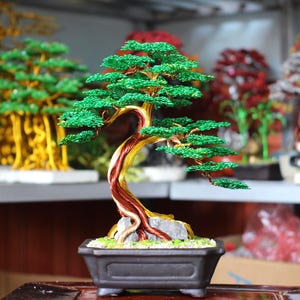 May include: A decorative wire bonsai tree sculpture featuring a twisted trunk with metallic copper and gold tones, topped with vibrant green wire foliage. The sculpture is anchored in a dark rectangular ceramic pot filled with small light-colored stones.