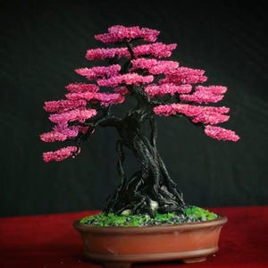 Op de afbeelding: Een handgemaakte bonsai-boomsculptuur met een donkere, getextureerde, gedraaide stam en takken versierd met felroze bladeren. De boom staat in een ondiepe, ovale bruine keramische pot gevuld met kleine grijze steentjes en groen mos.