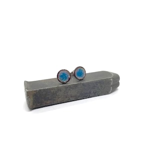 May include: A pair of small, round earrings with a blue and white enamel design. The earrings are set on a silver base and are displayed on a metal surface.