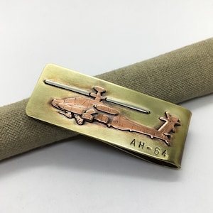 Peut inclure: Un porte-monnaie en laiton avec un hélicoptère gravé en cuivre. L'hélicoptère est un AH-64 Apache. Le porte-monnaie est de forme rectangulaire avec des bords arrondis.