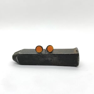 May include: A pair of small, round orange stud earrings with black outlines. The earrings are sitting on a metal surface.