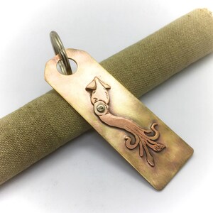 May include: A brass and copper key chain featuring a detailed squid design. The squid is depicted in a copper tone, while the rest of the keychain is brass.