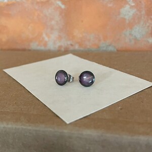 May include: A pair of small, round stud earrings with a purple and silver design. The earrings are on a white surface.