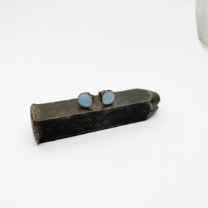 May include: A pair of small, round, light blue stud earrings with silver posts. The earrings are sitting on a metal surface.
