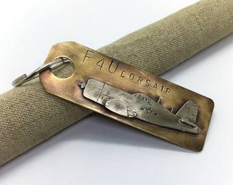 May include: A brass keychain with a silver F4U Corsair airplane engraved on it. The keychain has a silver key ring and the words "F4U Corsair" are engraved on the brass.