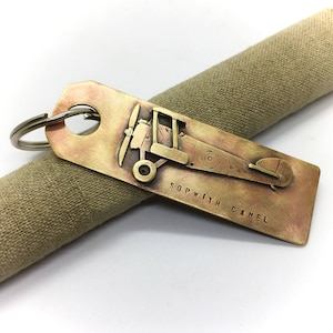 May include: Brass key chain with a detailed image of a Sopwith Camel airplane. The keychain has a ring for attaching keys and the text "Sopwith Camel" is engraved on the bottom.