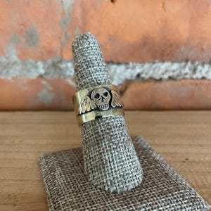 May include: A gold-toned ring with a skull and wings design. The ring is on a brown fabric ring holder.