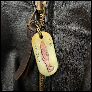 May include: A brass key chain featuring a P-51 Mustang aircraft design. The keychain is attached to a brown leather jacket zipper.