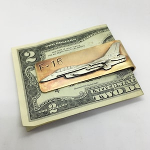 May include: A silver and copper money clip featuring a detailed F-16 fighter jet design. The clip is holding a two dollar bill.