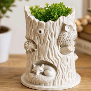 Op de afbeelding: Een crèmekleurige, cilindervormige plantenbak in de vorm van een boomstronk met gestructureerde schors. De buitenkant is voorzien van gebeeldhouwde figuren van een uil, een egel en een slapende vos. De opening bevat een kleine, groene kunstplant.