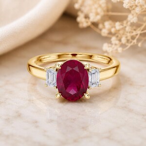 Op de afbeelding: Een gouden ring met een grote, ovaal geslepen, dieprode robijn in het midden, geflankeerd door twee kleinere, rechthoekige, baguette geslepen, helderwitte diamanten. De edelstenen zijn gezet in een gepolijste geelgouden ring met vier pootjes.