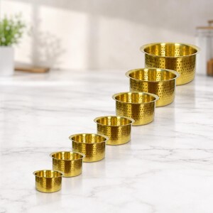May include: A set of seven graduated gold-toned metal bowls, each featuring a perforated design with small circular holes throughout the sides and base. The bowls are arranged in a diagonal line, decreasing in size from the largest to the smallest.