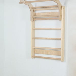 Peut inclure: Une échelle d'escalade murale en bois clair dotée de barreaux horizontaux et d'une barre de traction étendue sur la partie supérieure. La structure se compose de deux panneaux latéraux verticaux reliés par plusieurs barreaux cylindriques.