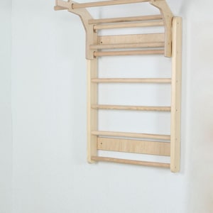 Puede incluir: Una escalera de escalada de madera montada en la pared que presenta una serie de peldaños horizontales y una sección superior de barra de dominadas. La estructura está hecha de madera natural clara con un acabado liso y dos rieles laterales.