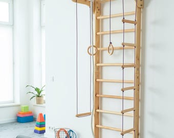 Könnte beinhalten: Ein hölzerner Kletterrahmen mit einer Leiter, Ringen und einer Schaukel. Der Rahmen ist an einer weißen Wand in einem Raum mit grauem Boden montiert. Eine zusammengerollte Trainingsmatte und andere Fitnessgeräte sind sichtbar.