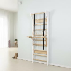 Puede incluir: Una espaldera de madera montada en la pared con un marco blanco y barras horizontales de color madera natural. La unidad incluye un accesorio de barra de dominadas desmontable y dos correas negras ajustables con anillas de gimnasia de madera.