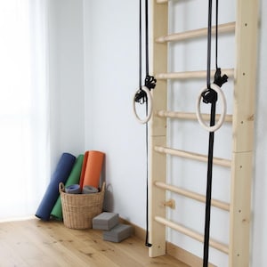 Puede incluir: Una escalera de pared de madera clara con pelda&ntilde;os horizontales y dos anillas de gimnasia de madera suspendidas por correas negras. Una cesta de mimbre tejida contiene esterillas de yoga enrolladas azules, verdes y naranjas, con dos bloques de espuma grises.