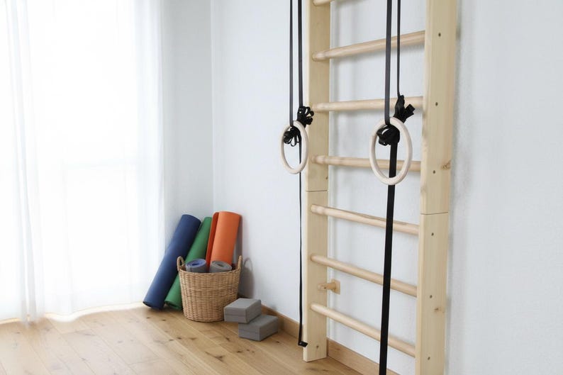 Puede incluir: Una escalera de pared de madera clara con pelda&ntilde;os horizontales y dos anillas de gimnasia de madera suspendidas por correas negras. Una cesta de mimbre tejida contiene esterillas de yoga enrolladas azules, verdes y naranjas, con dos bloques de espuma grises.