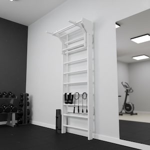 Op de afbeelding: Een wit houten wandgemonteerd klimrek met een optrekstang aan de bovenkant. De verticale ladder heeft meerdere horizontale sporten, met twee zwarte enkelbanden en twee zwarte springtouwen die aan het middengedeelte hangen.