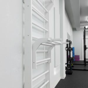 Puede incluir: Una espaldera blanca montada en la pared que presenta una serie de pelda&ntilde;os horizontales y un accesorio de barra de dominadas desmontable que se extiende hacia afuera. La estructura es de material blanco s&oacute;lido y est&aacute; fijada a la pared.